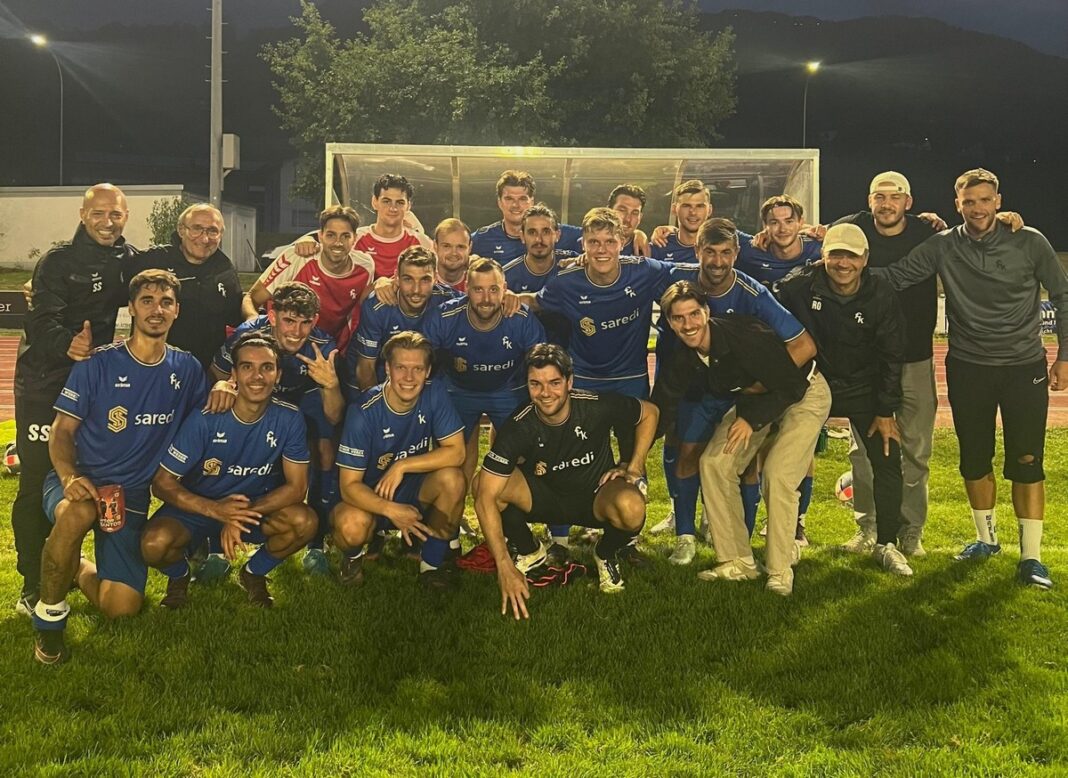 fckuessnacht Group of soccer players and staff posing for a team photo on a grass field at night, all wearing blue uniforms with a sponsor logo.