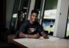 FC Luzern verpflichtet Faveurdi Bongeli leihweise Young man sits at a marble table, writing on papers with a red pencil, indoors near large windows.