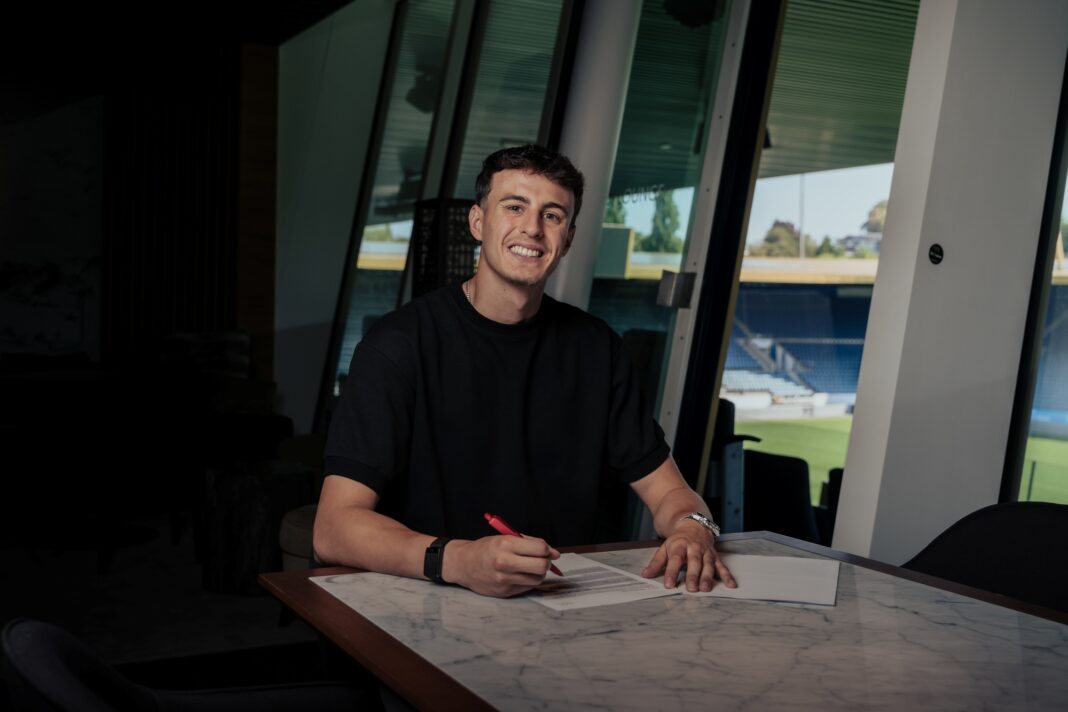 Man smiling while signing documents at a marble table inside a modern building, using a red pen.