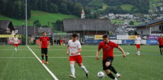 Soccer match on artificial turf; red and white team players chase the ball, with a player in red kicking the ball while a white-clad opponent closes in, hillside village in background