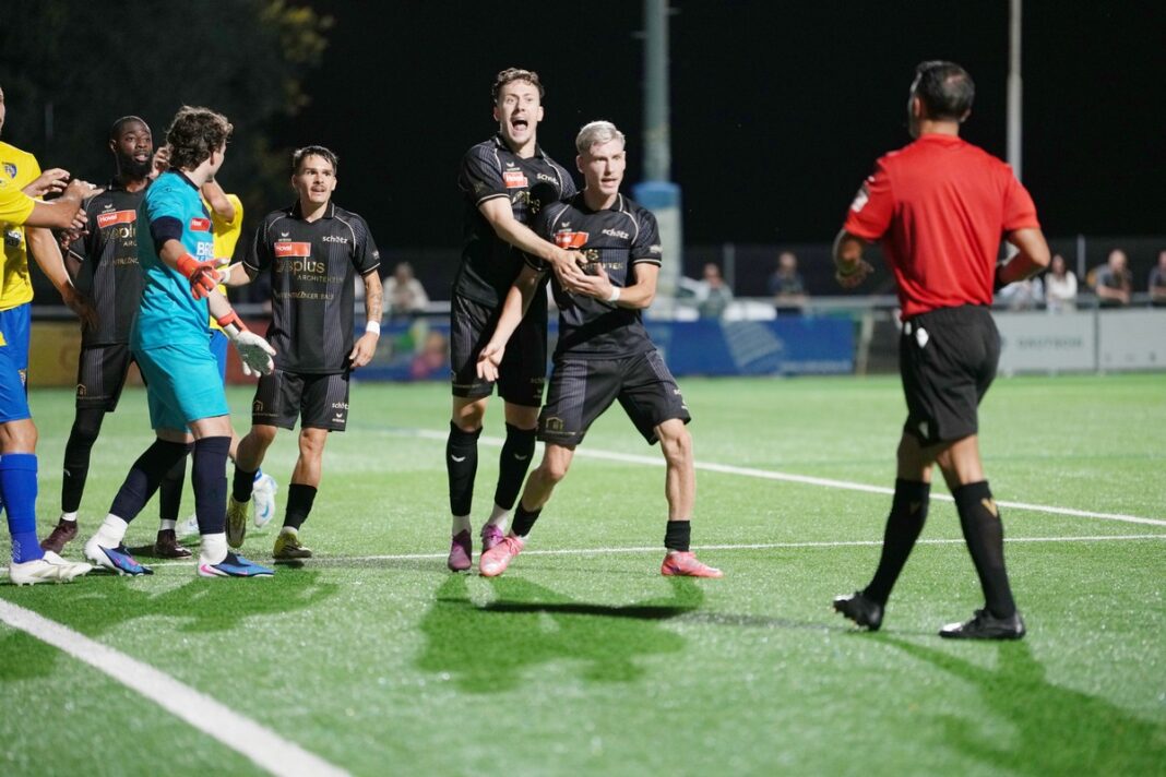 fcschoetz_fclangenthal14 Soccer players in black uniforms celebrate a goal on a bright green field at night, with a referee in red nearby.