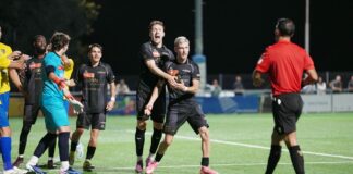 Soccer players in black uniforms celebrate a goal on a bright green field at night, with a referee in red nearby.