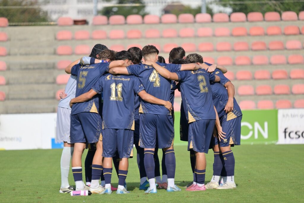 fcsolothurn_fcluzernu211-1024x683 Soccer players in blue uniforms huddle with arms around each other on a grass field in a stadium with red seats behind them