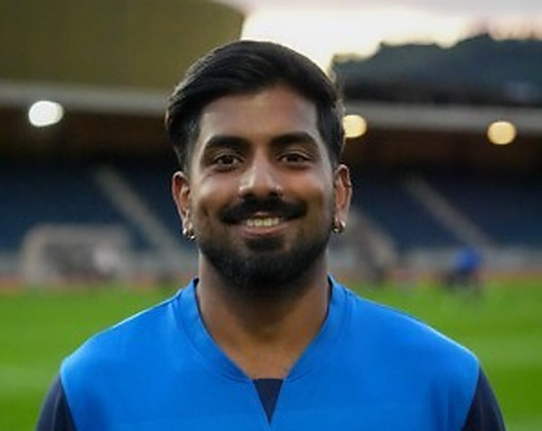 lsc_1200px Smiling man with dark hair and a beard wearing a blue sports jersey on a grassy field at a stadium.