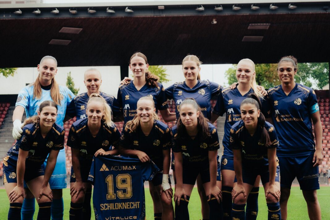 Group portrait of female soccer players in blue uniforms posing together on the field, stadium seating in the background, with player wearing jersey #19 Schildknecht in front.