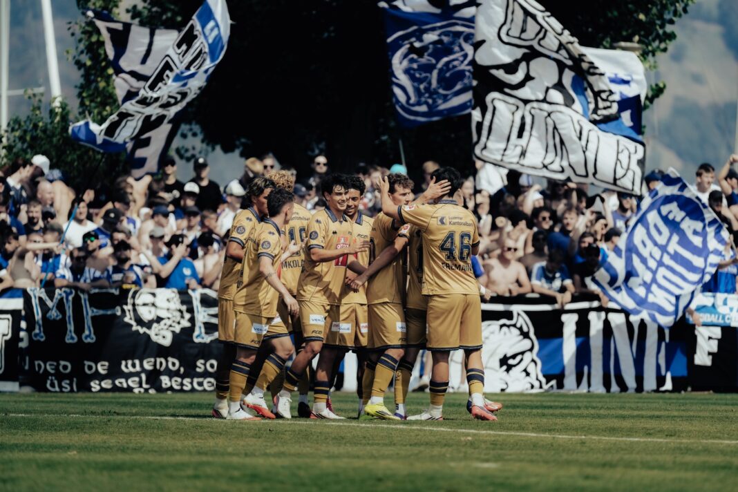 scbuochs-fcluzern-schweizer-cup-runde-1 Soccer players in gold uniforms celebrate on the field while fans wave blue and white flags in the stands in the background.