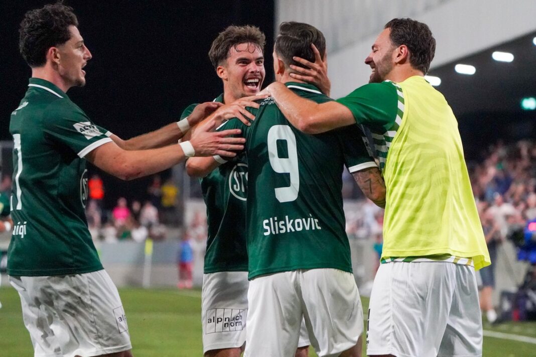 Soccer players in green celebrate a goal, hugging on the field.
