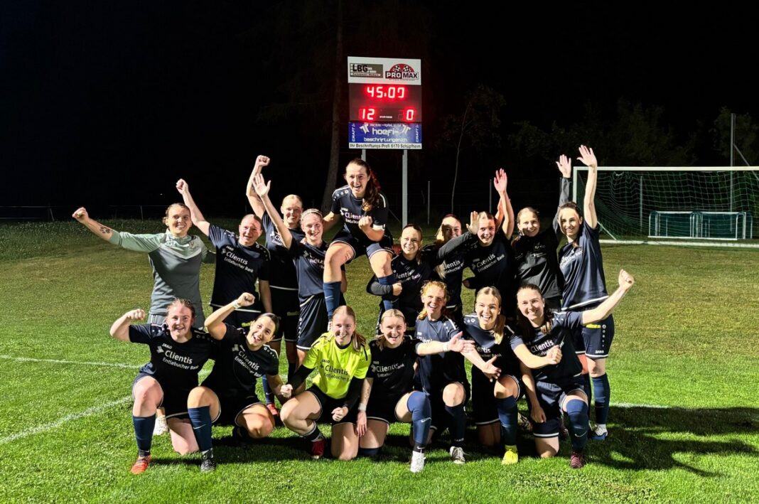 Group of female soccer players in dark uniforms celebrate on a lit grass field, one player lifted on teammates' shoulders.