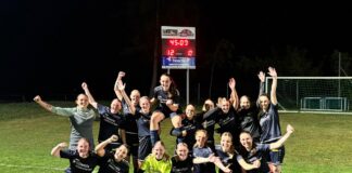 Group of female soccer players in dark uniforms celebrate on a lit grass field, one player lifted on teammates' shoulders.