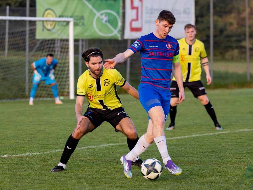 Davide-Di-Berardino-in-Blau-entkommt-Verteidiger-Enea-Bossart-1024x768 Soccer match: blue-clad player dribbles ball while yellow-jersey opponent tries to tackle, on a grassy field with goal in background.