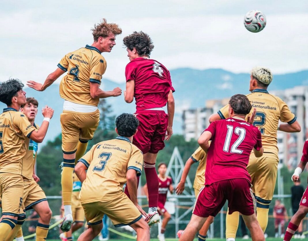 WhatsApp Image 2026-09-02 at 07.52.26 Soccer players in gold and maroon uniforms jump to head the ball during a match on an outdoor field.