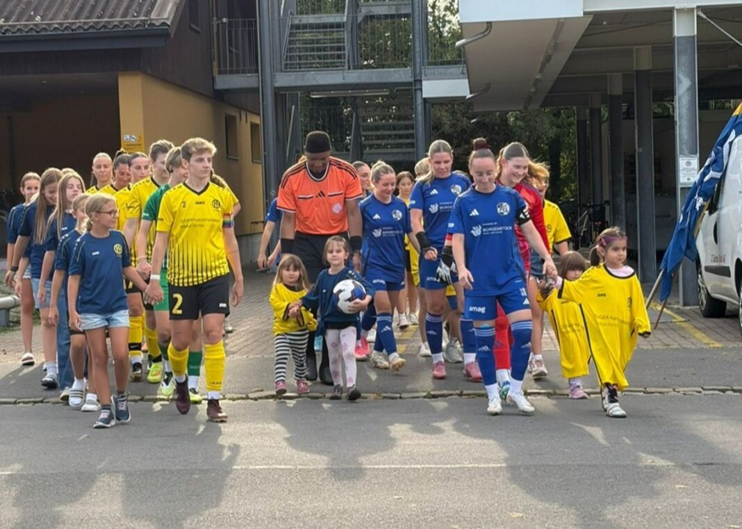 Line of young female soccer players in yellow, blue, and other uniforms walk in a line outdoors, some holding hands with small children at the front.
