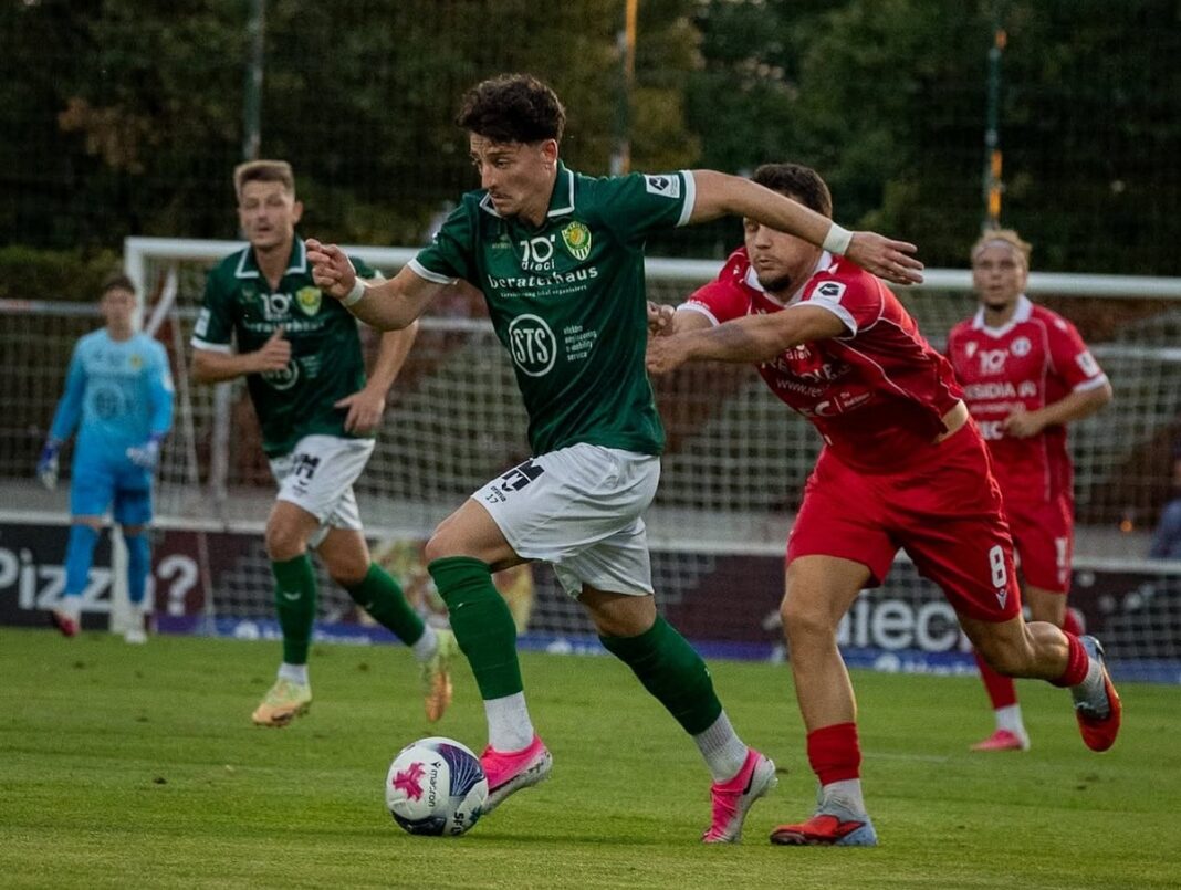 Two soccer players in green and red contest the ball on a grassy pitch, green player ahead with pink shoes leading the dribble as red defender reaches in.