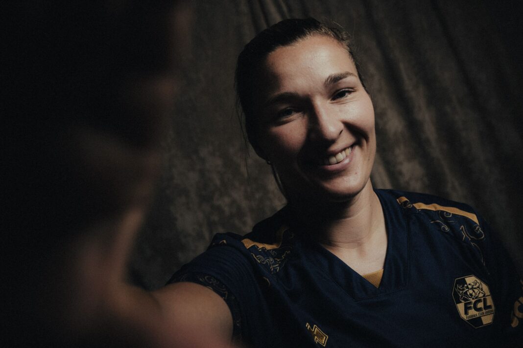 Close-up portrait of a smiling person wearing a navy blue sports jersey with a crest, against a dark textured background.